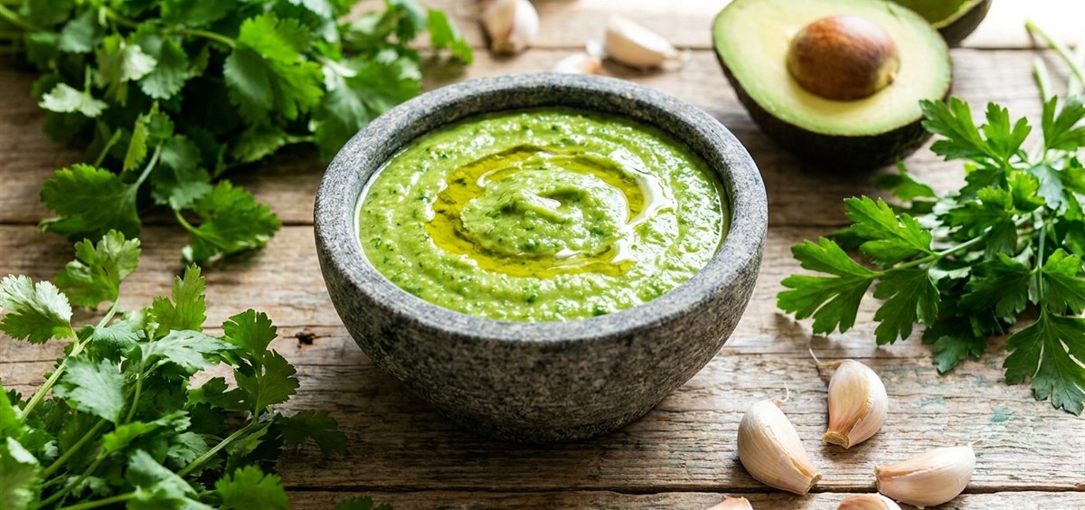 Stone bowl of fresh green Venezuelan guasacaca sauce surrounded by cilantro, parsley, avocado and garlic on a wooden table
