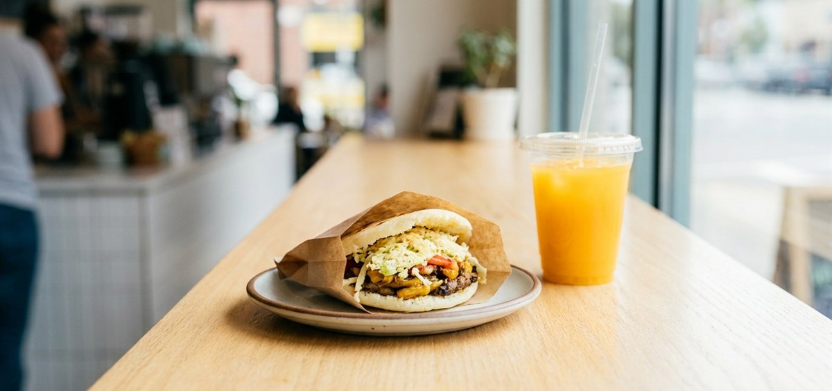 Filled Venezuelan arepa wrapped in paper with a to-go drink on a light cafe counter, ready for a quick lunch